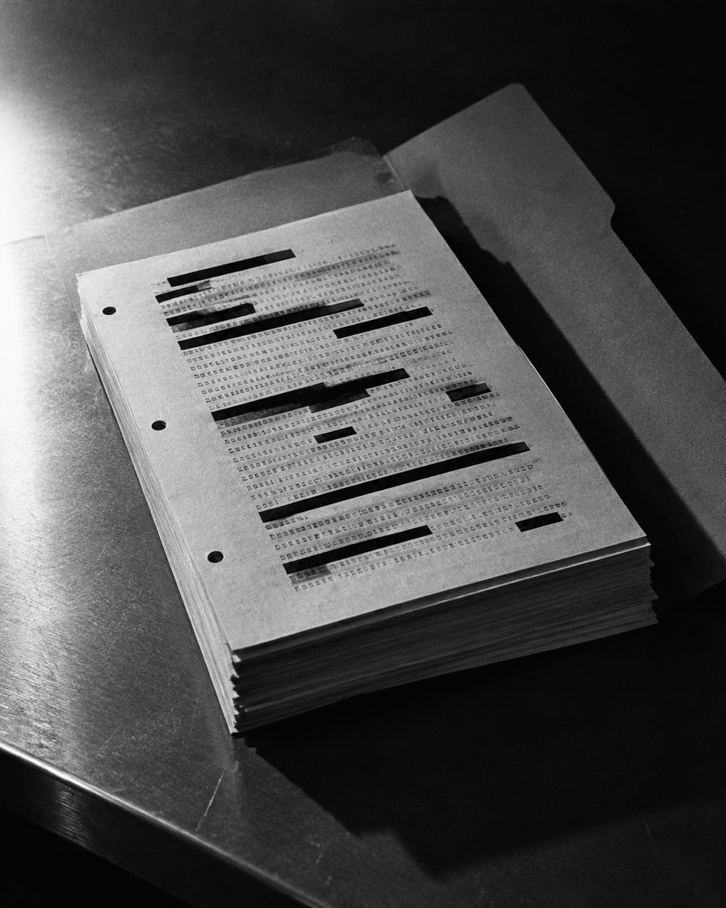 A stack of declassified documents bearing redaction bars, resting on a steel table